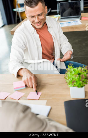 young businessman pointing at empty sticky note while sitting near colleague at workplace Stock Photo