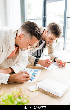 two young businessmen analyzing papers with graphs and charts while working on startup project together in office Stock Photo