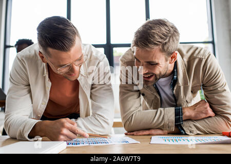 two young smiling businessmen analyzing papers with graphs and charts while working on startup project together in office Stock Photo