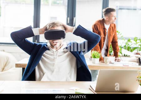 young businessman using vr headset and holding hands behind head while sitting at workplace in office Stock Photo