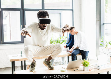 african american businessman using vr headset and squatting on desk while colleagues working in office Stock Photo
