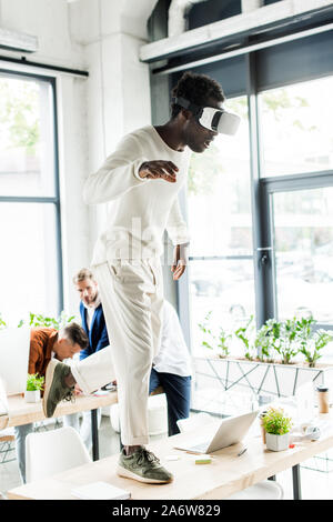 african american businessman using vr headset and standing on desk while colleagues working in office Stock Photo