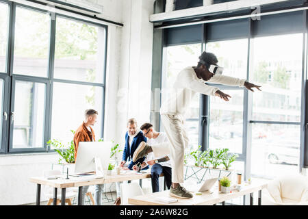 african american businessman using vr headset and standing on desk while colleagues working in office Stock Photo