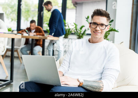 young businessman using laptop and smiling at camera while working near colleagues in office Stock Photo