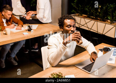 tired african american businessman drinking coffee to go while sitting at workplace at night in office near colleagues Stock Photo