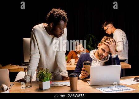 african american businessman standing near exhausted colleague holding coffee to go while working at night in office Stock Photo