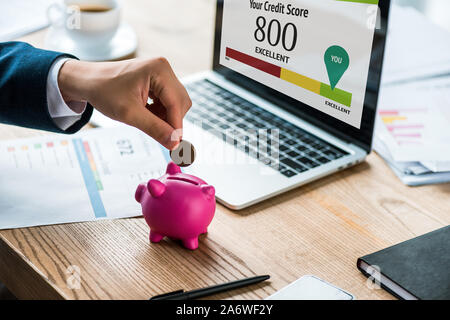 cropped view of businessman putting coin into pink piggy bank near laptop with your credit score letters Stock Photo