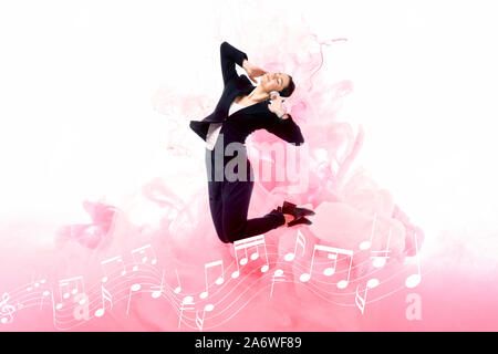 young businesswoman listening music in headphones while jumping on background with music notes and pink smoke splashes isolated on white Stock Photo
