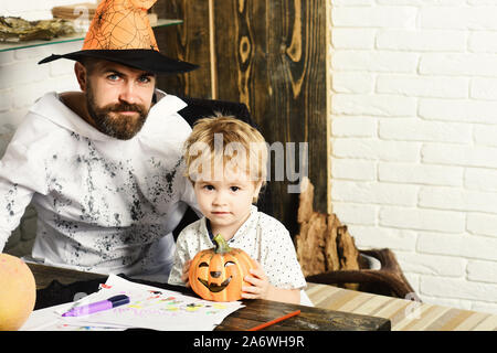 Guy and boy on wooden and white brick wall background. Witcher and little magician in Halloween room. Halloween and holiday concept. Man in orange hat and kid with smiling faces hold carved pumpkin Stock Photo