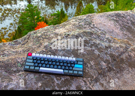 A mechanical computer keyboard with grey, white, black, red and blue keys seems a little out of place on the rock of a cliff above trees and a pond in Stock Photo