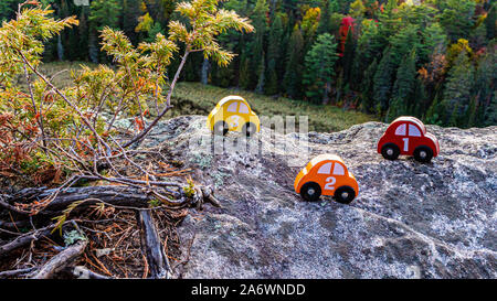 A series of red, orange and yellow toy cars, numbered 1, 2 and 3, sit on the rock of a cliff overlooking a forest. The wooden toys are placed next to Stock Photo