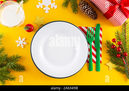 New year's table setting. Mock up white plate, knife, fork, glass with milk and christmas decorations on yellow background. Top view. Stock Photo