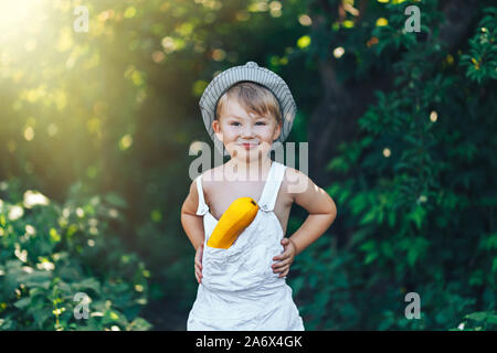 farmer kid with yellow squash in white casual clothes overalls and hat, standing in garden and smiling, harvest time. Stock Photo