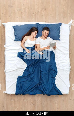 Bedtime. Young man and woman watching video on digital tablet cheerful lying under blanket on bed top view Stock Photo
