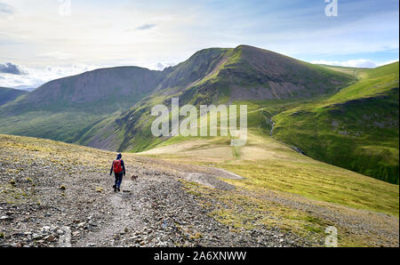 A hiker and their dog walking along a rugged path on a sunny day towards the summits of Crag Hill, Grasmoor and Sail in the Lake District, England, UK Stock Photo
