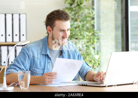 Casual happy entrepreneur working using laptop holding document comparing data at office Stock Photo