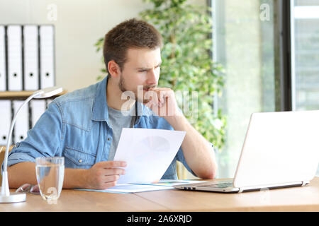 Pensive man holding document reading laptop content working at office Stock Photo