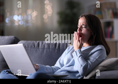 Fatigued woman using laptop yawning sitting on a couch in the night at home Stock Photo