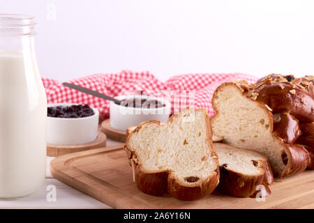 Still life with string of Christmas cake or sweet pastry with raisins and almond slices, glass of milk and bowls with fruit jam. Red kitchen towel on Stock Photo