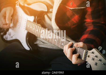 Stylish young musician in a beautiful plaid jacket plays powerful rock music on a black and white electric guitar, illuminated by bright light. Stock Photo