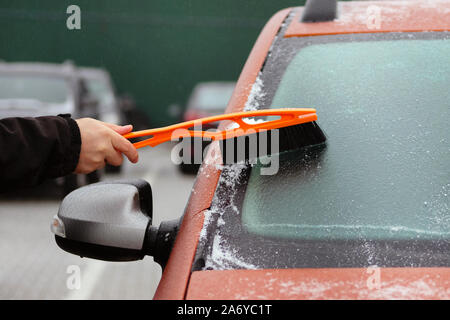 Brush in mans hand. Man clears snow from icy windshield of orange car. Man is brushing automobile window. Stock Photo