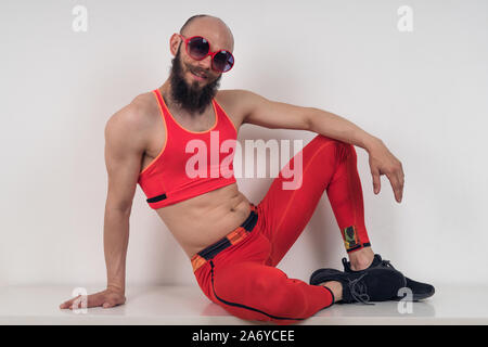 Funny guy in tight red tracksuit and sunglasses posing sitting. Stock Photo