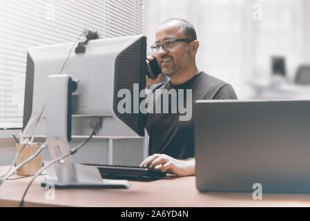 Smiling busy entrepreneur businessman working in the office talking using a mobile smartphone. Workplace and empty copy space for Editor's text. Stock Photo