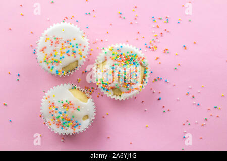 Three little white frosted white cupcakes in centre of pink background sprinkled with multi - coloured sprinkles on pink with copy space Stock Photo