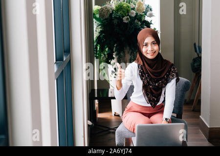 Happy Southeast Asian woman in hijab smiling and showing thumb up gesture while using laptop in office Stock Photo