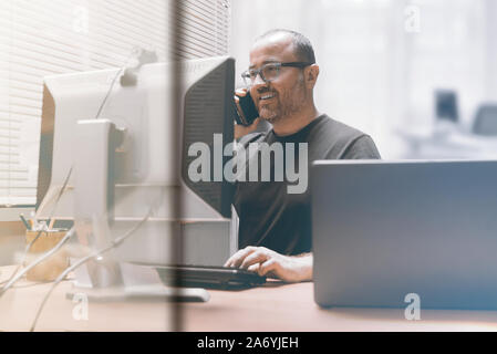 Smiling busy entrepreneur businessman working in the office talking using a mobile smartphone. Workplace and empty copy space for Editor's text. Stock Photo