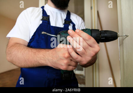 Construction worker and handyman works on renovation of apartment. Builder with electric drill drills nail hole into wall door at construction site. Stock Photo