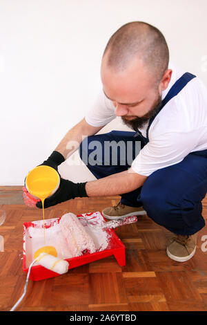 Construction worker doing finish renovation at apartment. Painter with black gloves pours yellow color from bucket into red bowl for paint mixing. Stock Photo