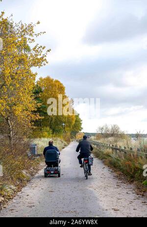 Two friends ride side-by-side, one on a bicycle and one on a mobility scooter, on the paved path along the seaside in Ystad, Sweden. Stock Photo