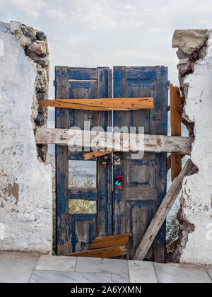 a derelict old weathered dilapidated door in door way wood wooden peeling paint held up by nailed planks of wood crumbling concrete white wall Oia Stock Photo