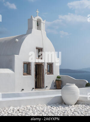 a white wash chapel church open door small bell tower with cross cruicifix blue sky cloud pot cobbles low wall foreground Oia Santorini Stock Photo