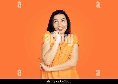 Caucasian young woman's portrait on orange studio background. Beautiful female brunette model in shirt. Concept of human emotions, facial expression, sales, ad. Copyspace. Dreamful, thoughtful. Stock Photo