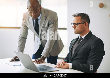 Two diverse businessmen discussing work together while using a laptop at a table in an office Stock Photo