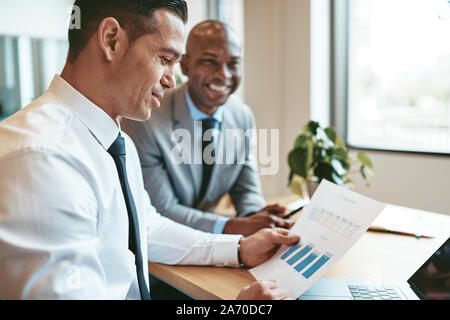 Two diverse businessmen smiling while going over charts and graphs on paper at a desk in an office Stock Photo