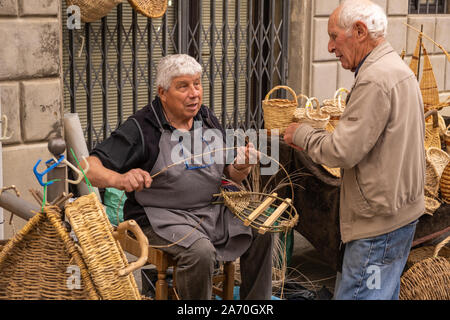 A male basket weaver at a craft market in a hilltop town in Italy talking to another old gentleman while making a basket Stock Photo