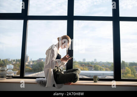 Blonde woman sitting on the window sill and texting boyfriend Stock Photo