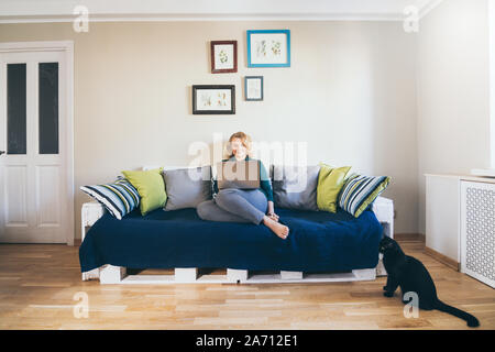 Young blonde Caucasian woman doing shoppings online on the couch at home, looking at laptop screen and smiling. Black cat on the foreground Stock Photo