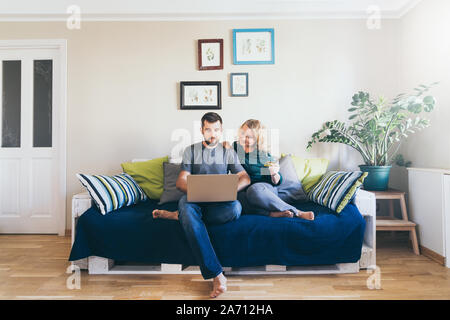 Young Caucasian couple doing shoppings online on the couch at home, looking at laptop screen and smiling Stock Photo