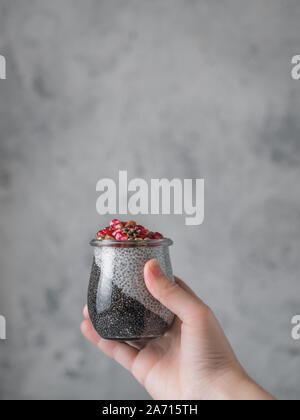 Chia pudding with black charcoal and white vanilla plant-based milk in woman hand on gray background. Female hand hold glass jar with chia puding served pomegranate and hemp. Copy space. Vertical Stock Photo