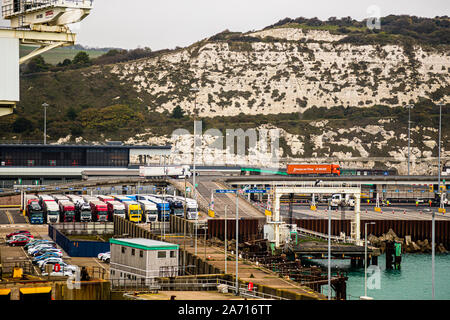 Lorries queue in several lanes for customs clearance at the ferry port of Dover, England, while a lorry bearing the slogan 'Always on Time' crosses a bridge above them. The town's White Cliffs form the backdrop. Little room remains to manoeuvre in the cramped harbour area when traffic comes to a standstill during checks. Stock Photo