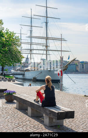 Young woman alone concept, rear view in summer of a young woman sitting alone on a bench in the Skeppsholmen waterfront, central Stockholm, Sweden. Stock Photo