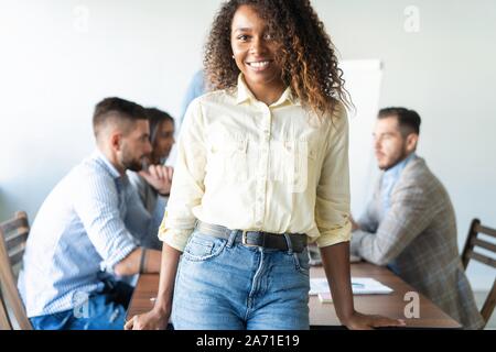 Beautiful young professional african woman standing in office while workers hold a meeting in background. Stock Photo