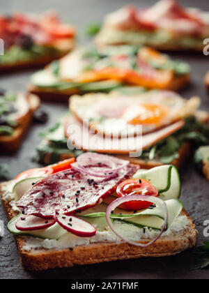 Close up view of different sandwiches with salami, vegetables and black sesame. Copy space for text. Assortment meat toasts on black background. Idea, creative concept for sausage maker. Vertical Stock Photo