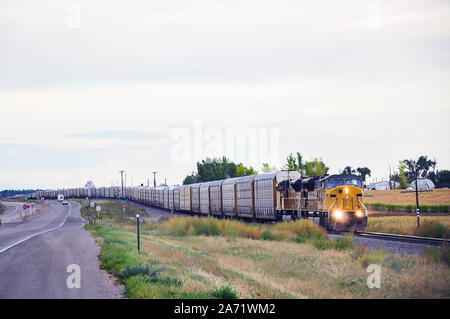 Long train hauling new cars down the track as vehichles stop at the crossing to let it pass. Stock Photo