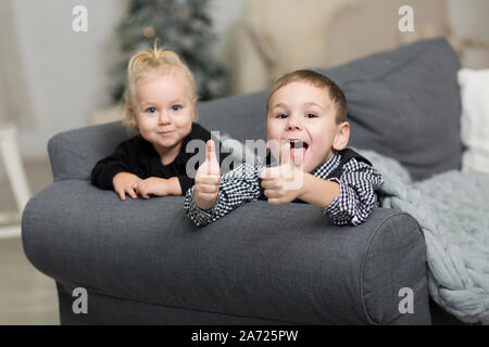 Little girl and boy lying t on the couch covered with a gray knitted blanket and smiling. Brother and sister grimacing and fooling around on the backg Stock Photo