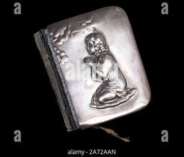 Miniature Edwardian-era Book of Common Prayer with a silver cover depicting a kneeling praying figure in relief, isolated on a black background Stock Photo